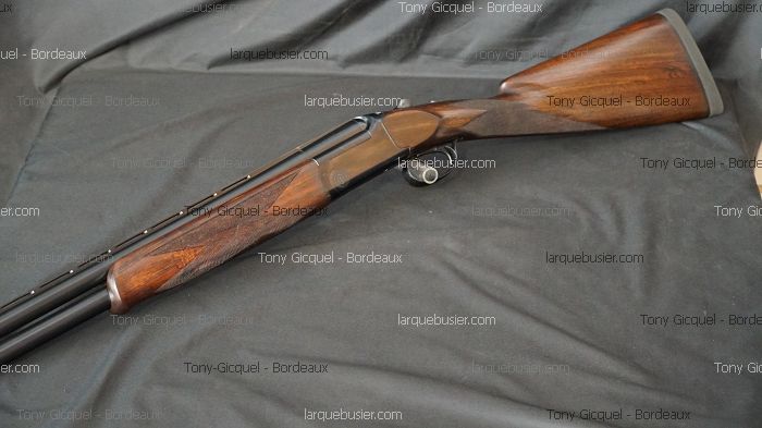 Fusil superposé élégant en bois foncé et canons noirs, reposant sur un tissu gris. Textes : Tony Gicquel - Bordeaux, larquebusier.com.