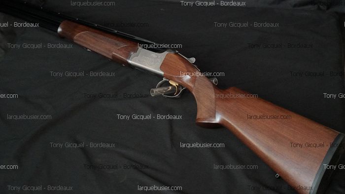 Fusil de chasse superposé avec crosse et devant en bois, bascule argentée et détente dorée, sur fond noir. Texte: Tony Gicquel - Bordeaux, larquebusier.com.