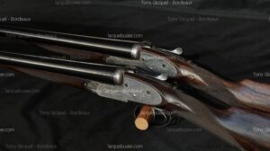 Deux fusils de chasse juxtaposés de luxe avec gravures fines et bois précieux, reposant sur support. Tony Gicquel - Bordeaux, larquebusier.com.