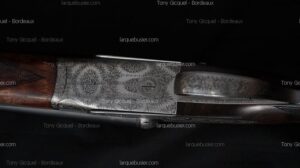 Culasse finement gravée d'une arme à feu vintage, motifs floraux et emblème circulaire, crosse bois foncé. Logos Tony Gicquel - Bordeaux, larquebusier.com.