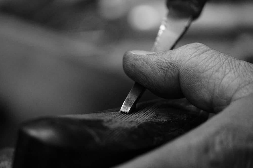 Main d'artisan en gros plan utilisant un burin pour graver un motif détaillé sur une surface texturée. Travail manuel précis en monochrome.