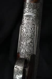 Gros plan d'un boîtier de fusil luxueusement gravé. L'argent brillant révèle des volutes florales, un dragon, un griffon et 'T GICQUE'.