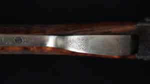 Récepteur de fusil de chasse en métal gravé de motifs floraux complexes, flanqué de bois. Surface polie reflétant la lumière.