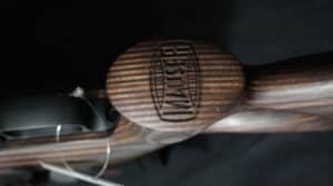 Gros plan sur la crosse en bois d'un fusil, avec le logo "MAUSER" gravé sur un médaillon circulaire et des rayures horizontales.
